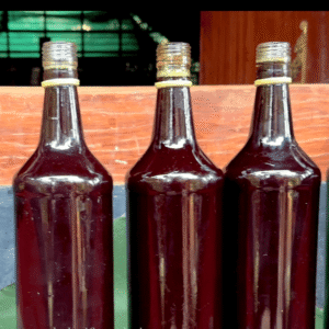 Close-up photograph of three clear glass bottles filled with dark reddish-brown Mukkoottu Tailam, a traditional Ayurvedic massage oil used in Kalaripayattu, lined up on a green surface (like a banana leaf) against a rustic wooden background.